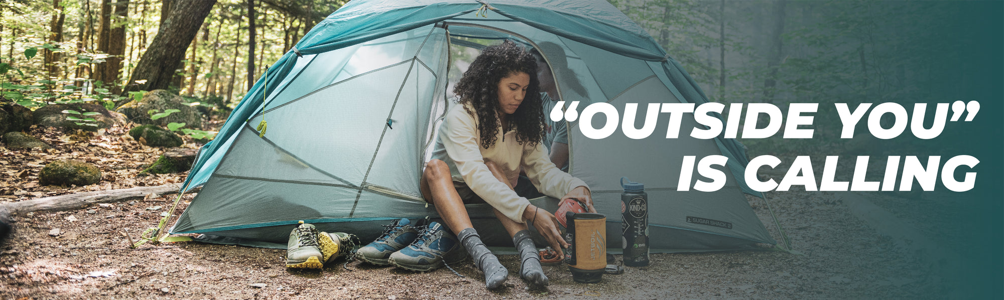Women in the woods in a camping tent with two pairs of shoes next to her wearing camping socks with text: "'Outside You' is Calling"